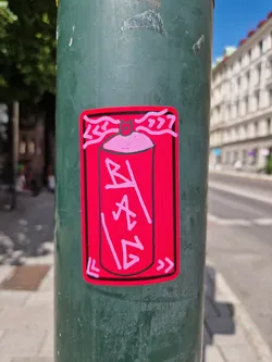 A pink sticker depicting a spray paint can with stylized graffiti lettering. The sticker has a rectangular shape with a slightly rounded top and bottom.  The main color is pink, with the lettering in a lighter pink. There are small decorative elements on either side of the main image. The sticker appears to be applied to a green metal pole.