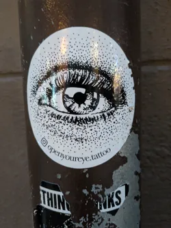 Street sticker The sticker is circular and predominantly white with black detailing. At its center is a detailed illustration of a human eye, focusing on the iris and eyelashes. The eye is rendered in black ink, with the iris showing concentric rings and a dark pupil. The eyelashes are sharp and expressive. The white background of the sticker is filled with a dense pattern of black dots, creating a textured or stippled effect that extends from the eye outwards towards the edges of the circle. Below the eye illustration, curved text is visible along the bottom edge of the sticker.