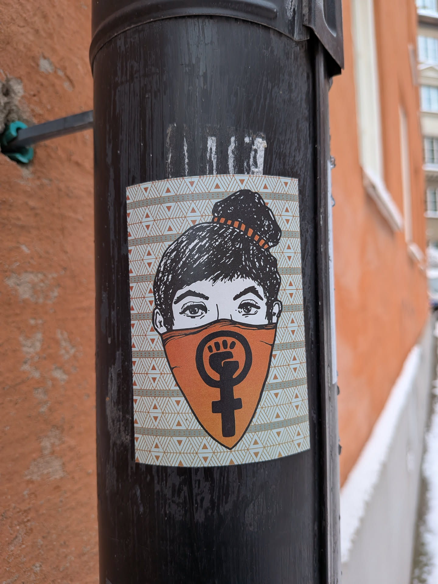 Street sticker The sticker features a stylized illustration of a woman's face, with dark hair pulled into a messy bun, secured by an orange hair tie. Her eyes are visible above an orange bandana-style mask that covers the lower half of her face. Centered on the mask is a bold black symbol combining a raised fist within a circle, superimposed on the Venus (female gender) symbol. The background of the sticker is a repeating geometric pattern of light blue/grey and white triangles. The overall color scheme is black, white, and orange, with accents of light blue/grey.