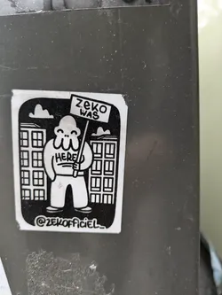 Street sticker Black and white sticker featuring a cartoon character holding a sign that says "ZeKO WAS HERE". The character is standing in front of buildings. The sticker has a simple design and uses bold outlines. At the bottom, there's an Instagram handle: @zeKOfficiel_
