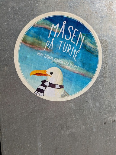 Street sticker This round sticker depicts a charming scene with a seagull as the focal point. The bird, adorned with a black and white striped scarf, stands out against a backdrop of a cloudy blue sky. The upper portion of the sticker features the text "Måsen På Turné" and "Våra näbbar klappar för blåvitt".