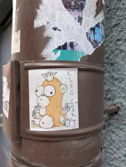 The sticker features a cartoonish, light orange monkey wearing a gold crown. It has large, wide eyes with black pupils and a small, white nose. The monkey is holding a glass filled with a frothy, light-colored beverage in its left hand, and its right hand is raised with two fingers extended in a peace sign. Its body is round and chubby, with fluffy white accents on its lower body and ears. The background of the sticker is white, and the outline and details are in black ink. To the right of the monkey, the words 'KING CHARLES' are written vertically in black, slightly stylized block letters.