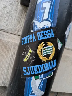 Street sticker A rectangular sticker with a black background, featuring two crest-like emblems and text in light blue block letters. On the left, a black shield with a crown on top contains a yellow diagonal banner with 'AIDS' in black text and '1891' below it, along with a sunburst design. On the right, a flag with horizontal green and white stripes displays 'H I V' vertically, encircled by a golden laurel wreath. Above the emblems, the text reads 'STOPPA DESSA', and below them, 'SJUKDOMAR'. The sticker appears slightly worn and wrinkled, affixed to a black pole over other blue and white stickers.