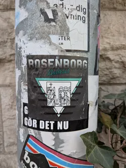 A rectangular sticker, predominantly black, featuring the name 'ROSENBORG' in bold, white, block letters at the top, with 'Ballklub' in a flowing, teal-green script directly beneath it. The central design is an inverted teal-green outlined triangle, within which a smaller, upright white triangle is framed. Inside this inner triangle, two figures are depicted in black line art: on the left, a cleric or bishop holding scales next to a church-like structure with a cross; on the right, a king or ruler also holding scales next to a castle tower. Below this emblem, the phrase 'GÖR DET NU' is written in bold, white, sans-serif capital letters. The sticker shows signs of wear and minor scuffs, consistent with outdoor exposure.