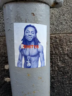 Street sticker A rectangular sticker featuring a blue-tinted grayscale portrait of rapper Lil Wayne from the chest up. He is shirtless, showcasing numerous tattoos across his chest and arms, and wears a prominent silver chain with a money bag pendant. Lil Wayne is smiling broadly, revealing his dental grills, and has his signature dreadlocks. The word 'KEHITYS' is emblazoned across his chest in large, bold orange letters with a white outline, with the 'K' being slightly larger and more stylized. The sticker is adhered to a textured, peeling grey metal pipe.