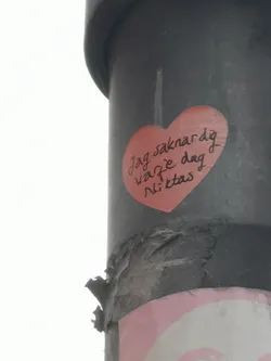 A vibrant red, heart-shaped sticker is prominently displayed on a dark, textured pole. The sticker has a slightly worn appearance with minor creases. Black, handwritten text in a casual, cursive style is inscribed across the center of the heart. The background of the image shows a bright, overcast sky.