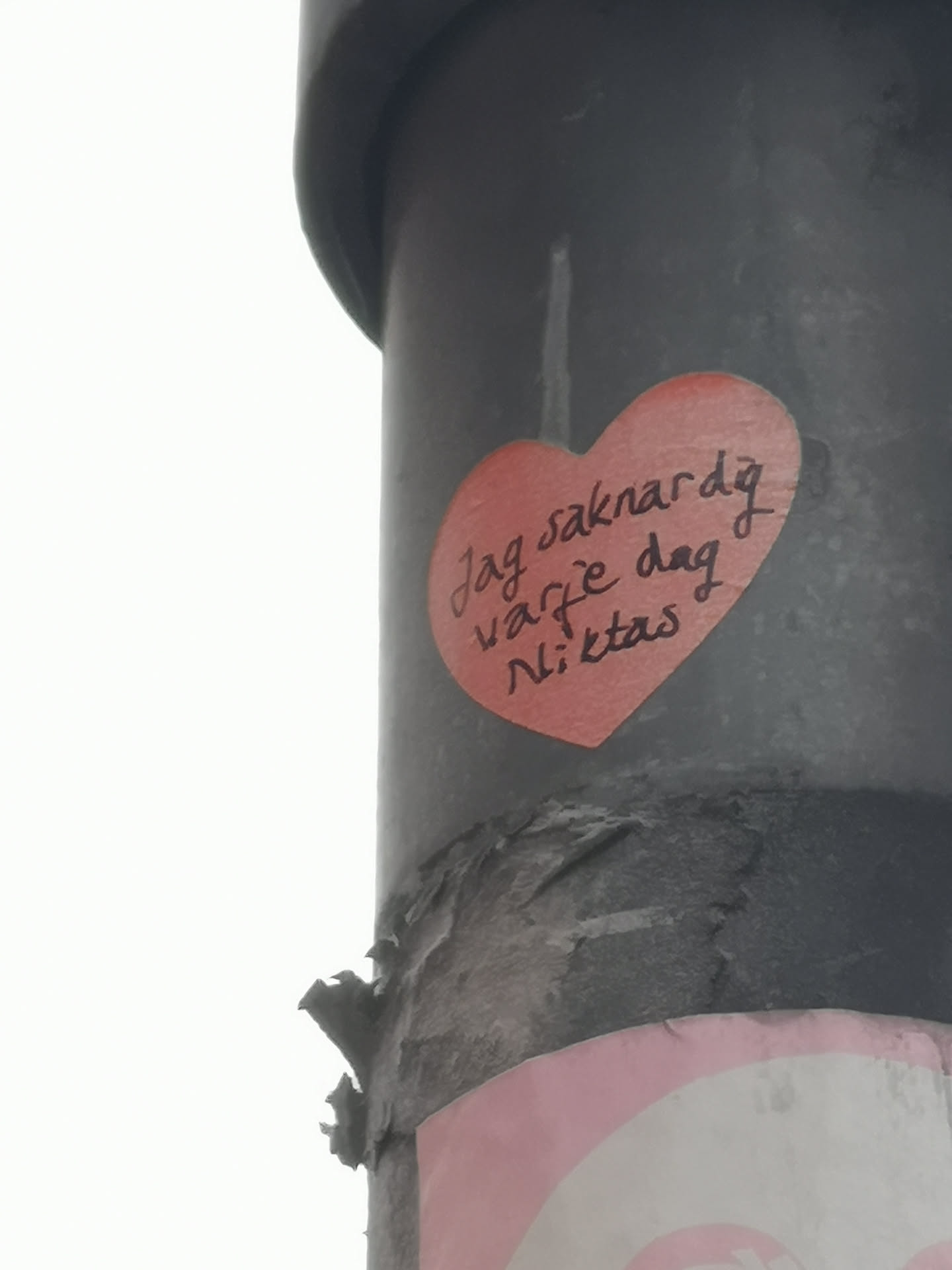 A vibrant red, heart-shaped sticker is prominently displayed on a dark, textured pole. The sticker has a slightly worn appearance with minor creases. Black, handwritten text in a casual, cursive style is inscribed across the center of the heart. The background of the image shows a bright, overcast sky.