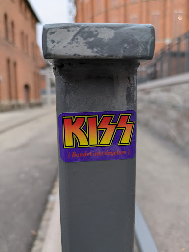 Street sticker A sticker featuring the iconic KISS logo is affixed to a gray metal pole. Below the logo, smaller text reads '(Bandet inte drycken)', which translates to '(The band, not the drink)' in Swedish.