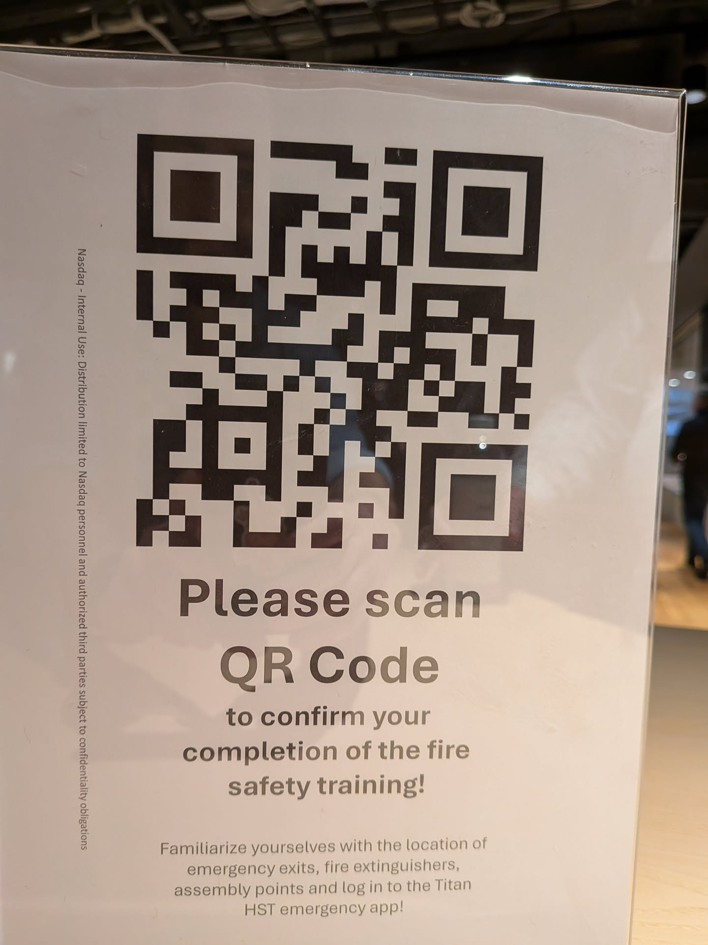The sticker is a white rectangular sheet with a large black QR code prominently displayed in the center. Below the QR code, there is text instructing the user to 'Please scan QR Code to confirm your completion of the fire safety training!'. Further down, smaller text provides additional information about familiarizing oneself with emergency exits, fire extinguishers, assembly points, and logging into the 'Titan HST emergency app'. Along the left edge of the sticker, there is vertical text indicating 'Nasdaq - Internal Use: Distribution limited to Nasdaq personnel and authorized third parties subject to confidentiality obligations'.