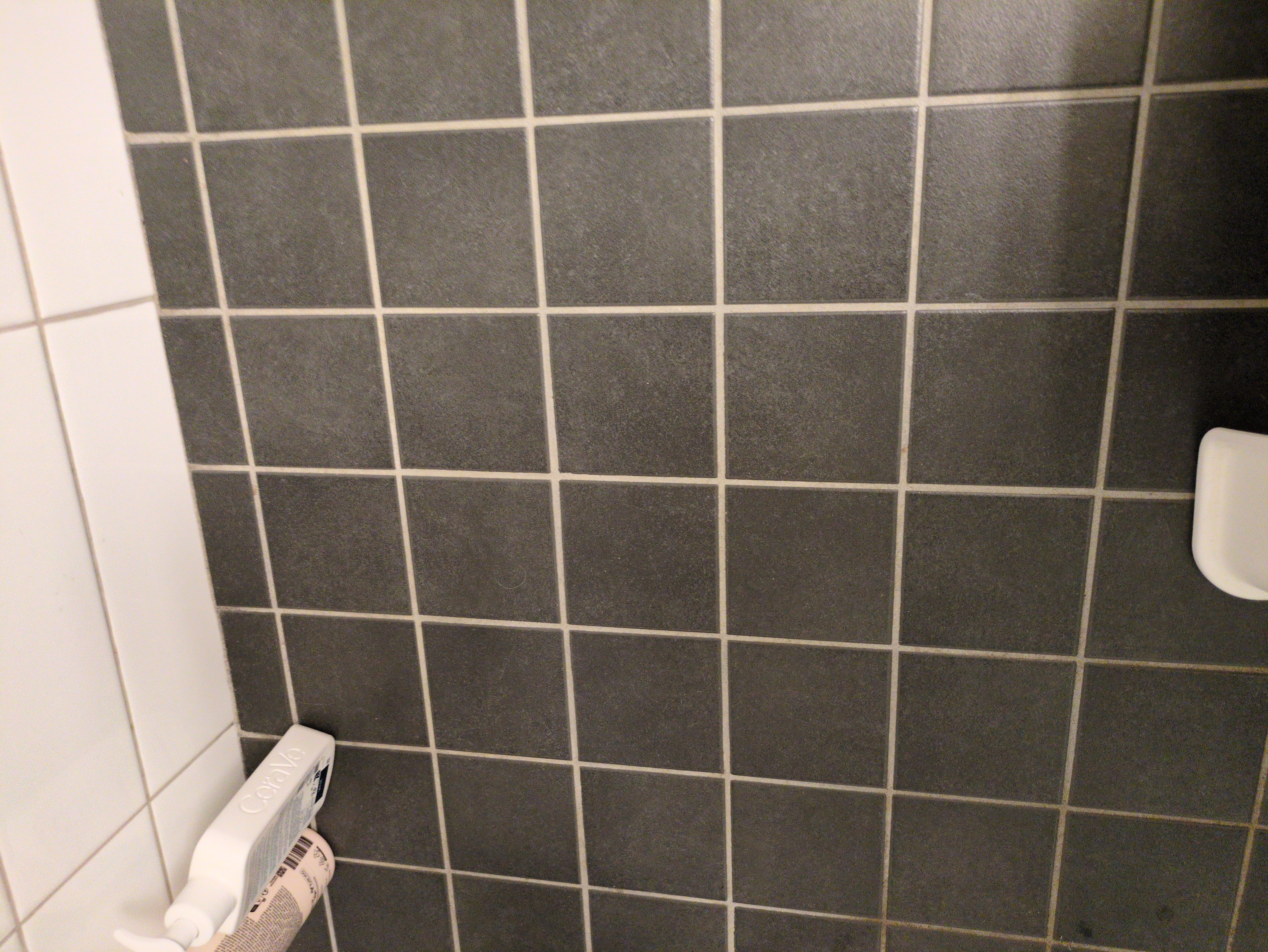 A photo of a bathroom wall covered in gray tiles. In the lower left corner, a white Cerave product is visible. The wall is simple and functional, typical of bathrooms.