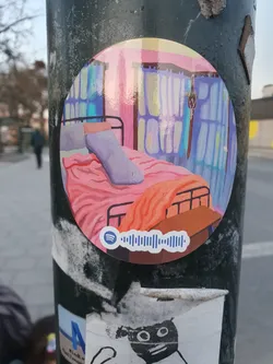 A round sticker featuring an illustration of a bedroom with a bed, blanket, and curtains. The style is reminiscent of  modern art. A Spotify code is included at the bottom.