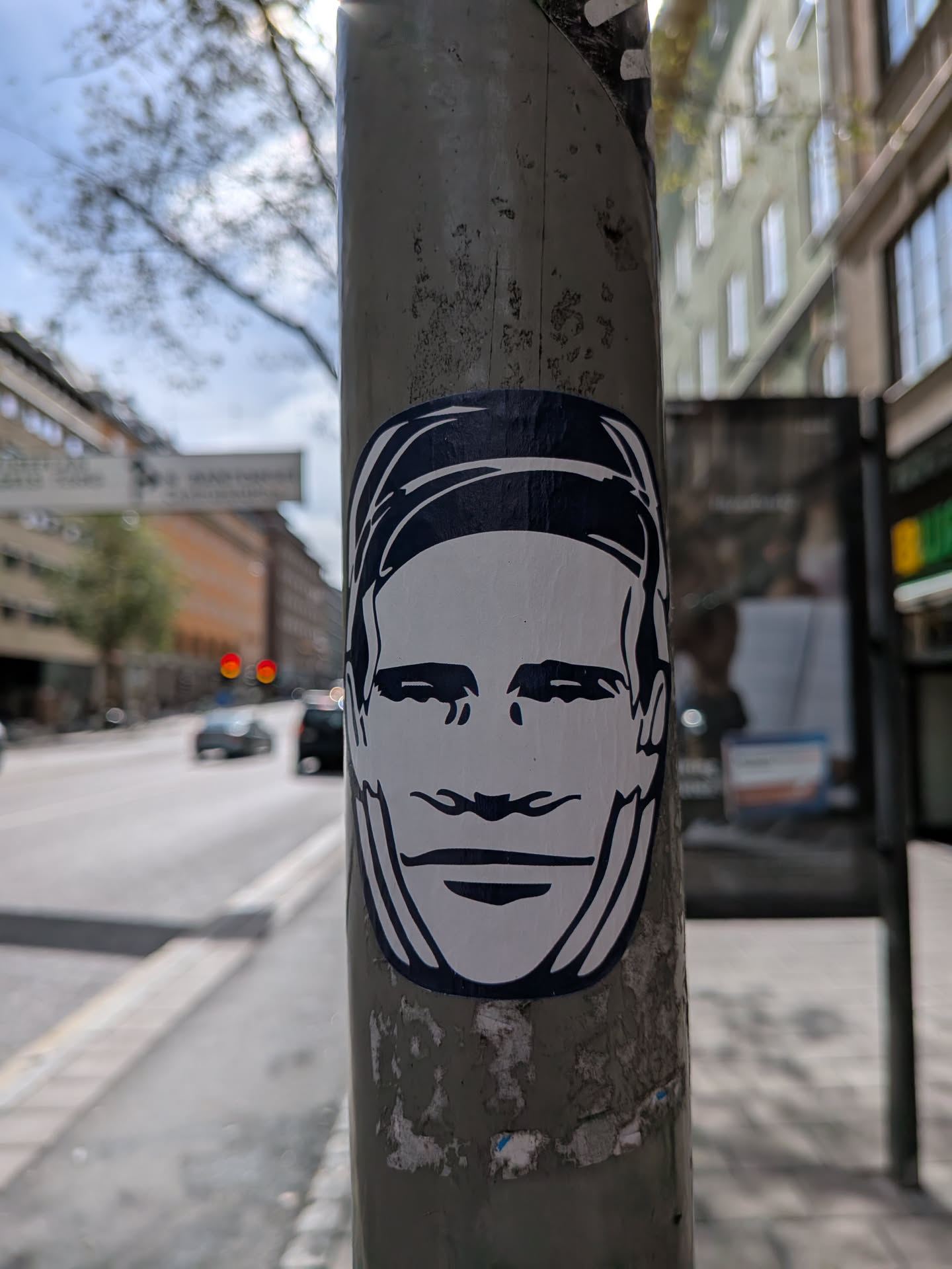 The sticker is a black and white, minimalist portrait of a face. The face is depicted with bold outlines and solid black areas for hair, eyebrows, eyes, and mouth. The expression appears neutral or slightly smiling. The sticker has a white background with black linework, and it is applied to a weathered grey pole.