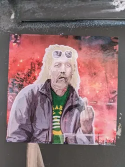 Street sticker This sticker depicts a man with long blond hair and sunglasses, wearing a gray jacket and a green shirt. He is giving the middle finger. The background shows a vibrant scene from a soccer match, possibly featuring flares or pyrotechnics. The style is bold and expressive.