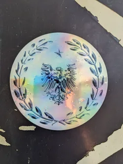 Street sticker This eye-catching holographic sticker showcases an intricate design. A double-headed eagle, symbolic of power and authority, takes center stage, surrounded by olive branches representing peace and victory. The holographic effect creates a mesmerizing iridescent shimmer.