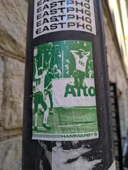 Street sticker A rectangular sticker, predominantly green and white, depicting a dynamic scene from a football (soccer) match or celebration. On the left, a player in white with green details raises their arms in triumph. On the right, another figure, also in white with green accents, stands with hands behind their head, displaying '1X2' on their chest. The background features a green field with white lines, suggesting a football pitch, and blurred figures of spectators or other players. A prominent white band across the middle features the word 'Alto' in bold green text. At the bottom, a thin white strip contains 'HAMMARBY' in green, followed by a small green circular logo. The sticker shows some wear and tear, with slight fading and minor damage along its edges.