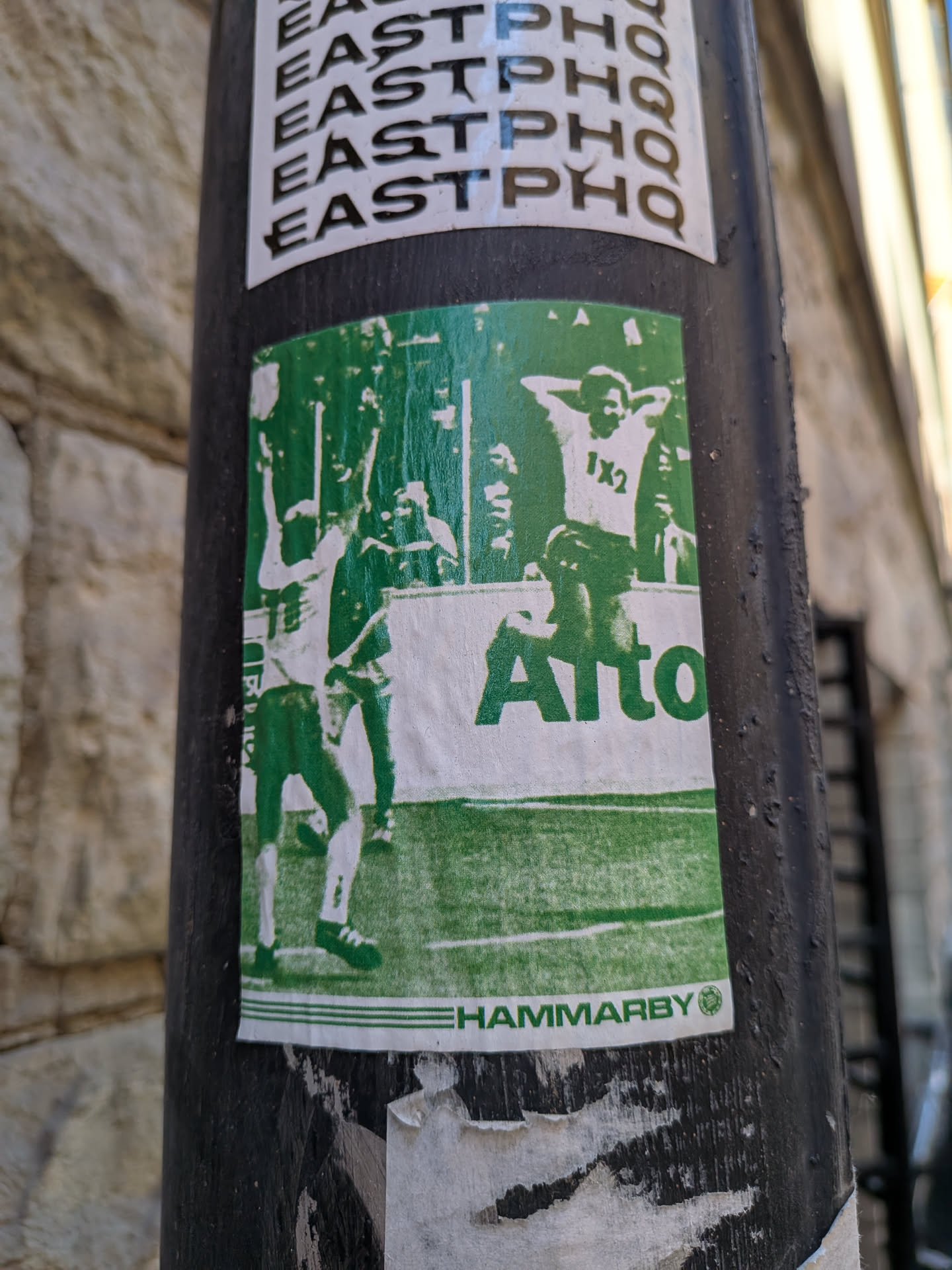 A rectangular sticker, predominantly green and white, depicting a dynamic scene from a football (soccer) match or celebration. On the left, a player in white with green details raises their arms in triumph. On the right, another figure, also in white with green accents, stands with hands behind their head, displaying '1X2' on their chest. The background features a green field with white lines, suggesting a football pitch, and blurred figures of spectators or other players. A prominent white band across the middle features the word 'Alto' in bold green text. At the bottom, a thin white strip contains 'HAMMARBY' in green, followed by a small green circular logo. The sticker shows some wear and tear, with slight fading and minor damage along its edges.