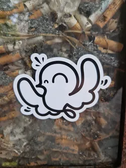 A small, white sticker featuring a simple black line drawing of a bird. The bird is depicted in a cheerful manner, with a slight smile and its wings outstretched. The sticker is applied to the glass of a public ashtray overflowing with cigarette butts.