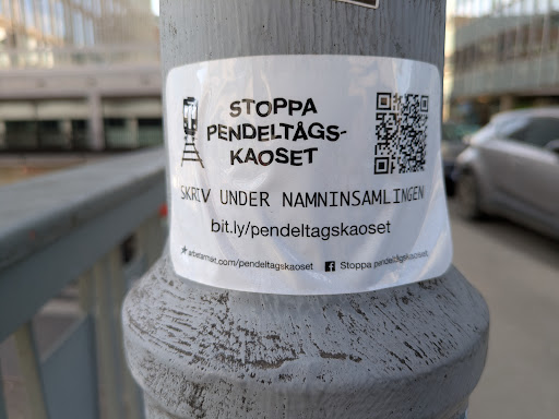 A sticker on a pole promoting a petition to stop commuter train chaos. The sticker features an image of a commuter train, a QR code, and text encouraging people to sign the petition. It also includes social media links.