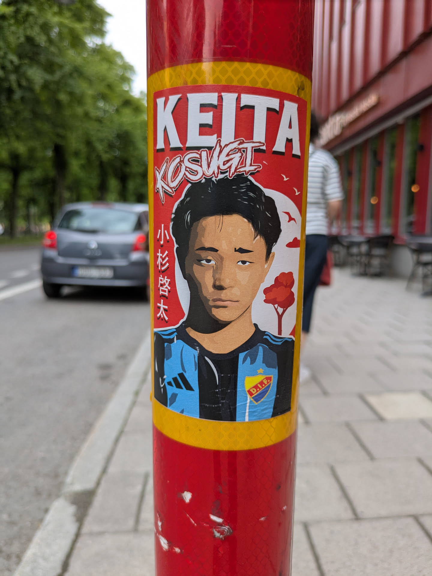 This is a portrait sticker on a red pole. The sticker has a drawing of Keita Kosugi, a football player. The text above the portrait says "KEITA KOSUGI" and there is Japanese text on the side. The player is wearing a blue and black jersey with the D.I.F. logo.