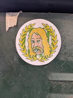 Street sticker The sticker is circular, featuring a cartoonish illustration of a man's head and upper chest. He has long, wavy blonde hair and a full, scruffy beard, both colored yellow with prominent black outlines. His skin is a light peach tone, and he has heavy-lidded, tired-looking eyes. A lit cigarette is depicted in his mouth. The man's head is framed by a green laurel wreath with yellow leaves, also outlined in black, giving it a regal yet disheveled appearance. The background within the wreath is a light off-white color. The overall style is reminiscent of a hand-drawn or marker-based illustration, with a somewhat rough and expressive quality.