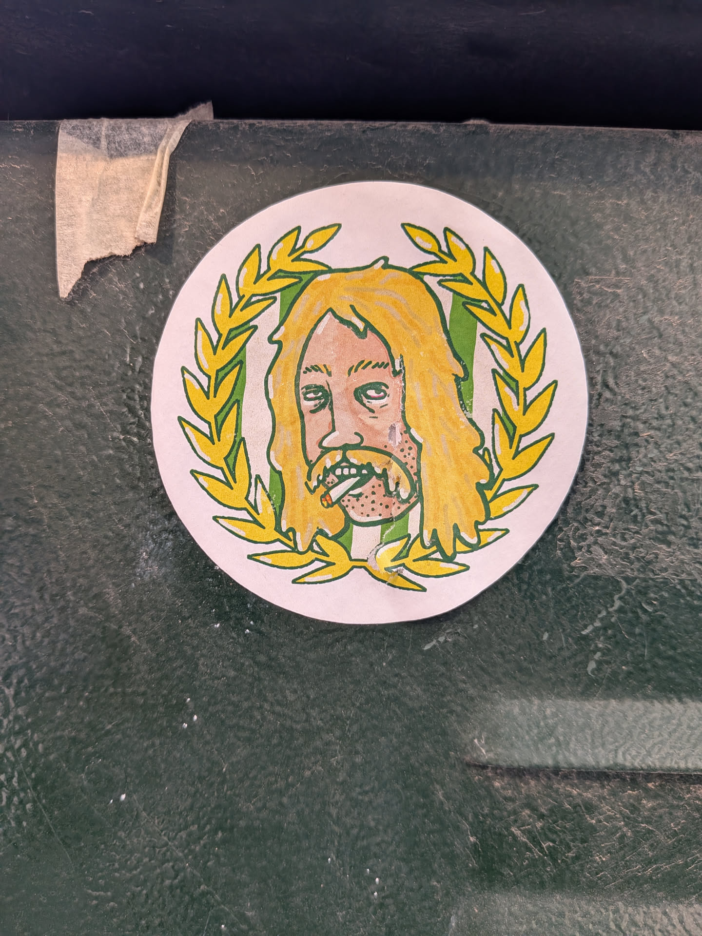 The sticker is circular, featuring a cartoonish illustration of a man's head and upper chest. He has long, wavy blonde hair and a full, scruffy beard, both colored yellow with prominent black outlines. His skin is a light peach tone, and he has heavy-lidded, tired-looking eyes. A lit cigarette is depicted in his mouth. The man's head is framed by a green laurel wreath with yellow leaves, also outlined in black, giving it a regal yet disheveled appearance. The background within the wreath is a light off-white color. The overall style is reminiscent of a hand-drawn or marker-based illustration, with a somewhat rough and expressive quality.