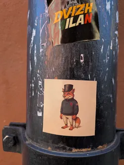 Street sticker The sticker features a full-body illustration of an anthropomorphic fox standing upright. The fox has reddish-brown fur, a white muzzle, and a friendly, slightly mischievous smile. It is dressed in a black top hat, a dark teal or forest green zippered sweater, and light beige trousers. On the left chest of the sweater, there's a red diamond-shaped emblem with a white symbol inside, possibly a stylized letter or logo. On the right sleeve, a small American flag patch is visible. The fox's bare feet and bushy tail are also visible. The background of the sticker is a plain, light cream or beige color, providing a clean contrast to the detailed illustration. The sticker itself is square with slightly rounded corners and appears to be made of paper or vinyl, adhered to a dark, weathered pole.