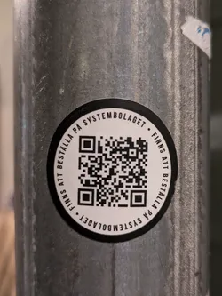 Street sticker A black and white circular sticker affixed to a grey metal pole. The sticker features a QR code in the center and text that reads: "FINNS ATT BESTÄLLA PÅ SYSTEMBOLAGET • FINNS ATT BESTÄLLA PÅ SYSTEMBOLAGET" which translates to: "Available to order at Systembolaget • Available to order at Systembolaget".