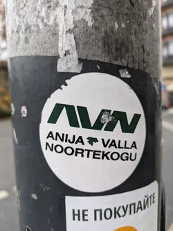 Street sticker A round sticker featuring the acronym AVN, which stands for Anija Valla Noortekogu (Anija Parish Youth Council). The sticker is white with dark green lettering and a small map-like symbol. It is stuck on a dark-colored pole.