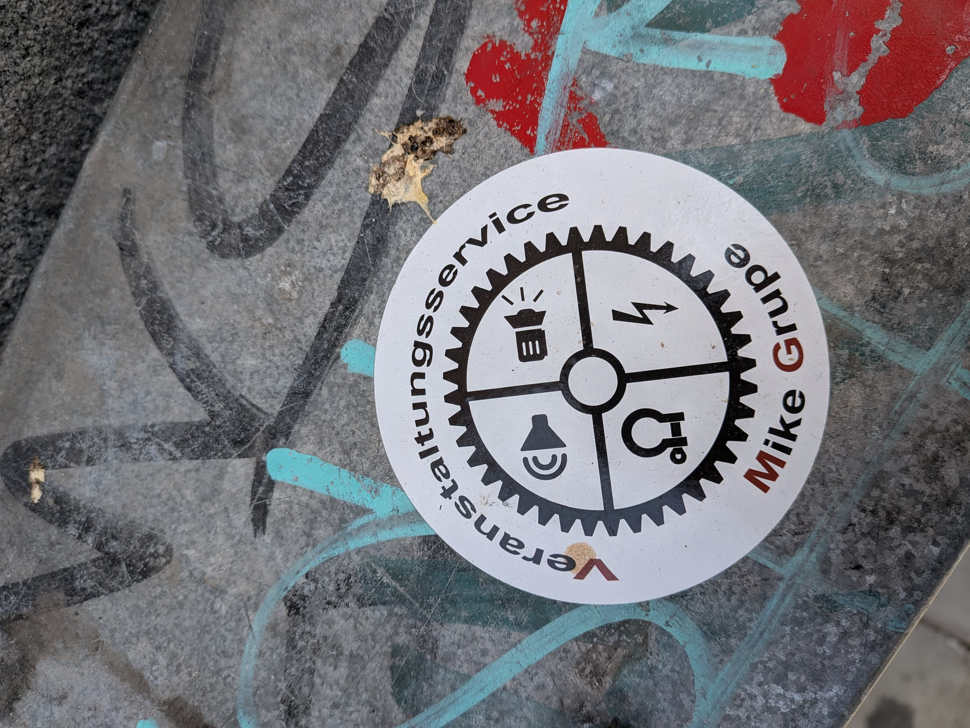 A circular white sticker featuring a black gear divided into four quadrants, each containing an event technology icon: a spotlight, a lightning bolt, a speaker, and a rigging clamp. The German text 'Veranstaltungsservice Mike Grupe' wraps around the gear with red initial letters.