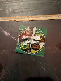 Street sticker The sticker is circular with a green background. The top section features a stylized illustration of a pilot wearing goggles and a helmet, with other figures in the background. Below the pilot, the text 'HAMMARBY' is prominently displayed in a bold, white, blocky font reminiscent of the Grand Theft Auto logo. Beneath this, 'SÖDERMALM' is written in a similar style. The bottom left quadrant contains a circular emblem with green and white stripes, surrounded by a yellow laurel wreath. The bottom right quadrant depicts a vintage-style football with a brown and white pattern, set against a yellow and green background with abstract black elements.