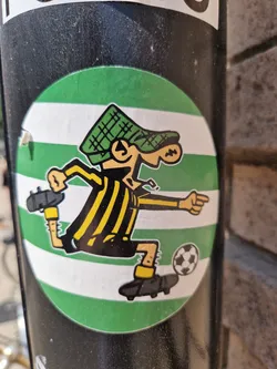 A round sticker featuring a cartoon drawing of a player kicking a soccer ball. The player is depicted with yellow and black striped shorts, a green hat, and is in a dynamic pose. The background of the sticker is green with white stripes.