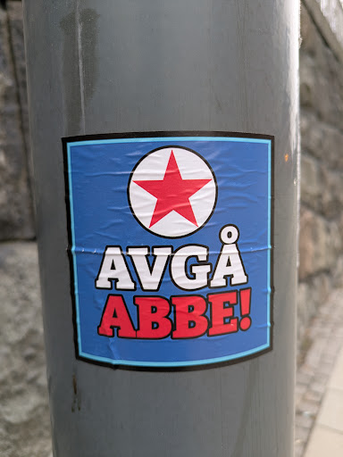 Street sticker A sticker with a blue background and a white circle containing a red star at the top. Below the star, the text "Avgå" is written in white, and "Abbe!" is written in red. The sticker is affixed to a gray pole.