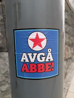 Street sticker A sticker with a blue background and a white circle containing a red star at the top. Below the star, the text "Avgå" is written in white, and "Abbe!" is written in red. The sticker is affixed to a gray pole.