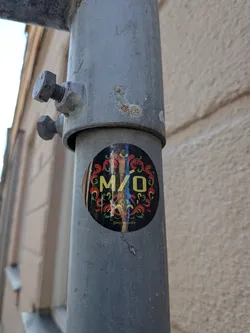 Oval black sticker on a metal pole. Features yellow 'M/Ó' text centered within red and green traditional folk-style floral ornaments. Small yellow text '@miomusikk' is at the bottom.