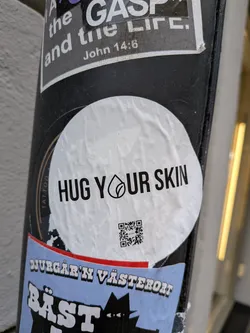 Street sticker A circular white sticker with bold black text that reads 'HUG YOUR SKIN'. The 'O' in 'YOUR' is replaced by a stylized black logo depicting a drop shape with a leaf inside. Below the text, there is a black and white QR code. The sticker appears slightly wrinkled, indicating its physical presence on a dark pole.