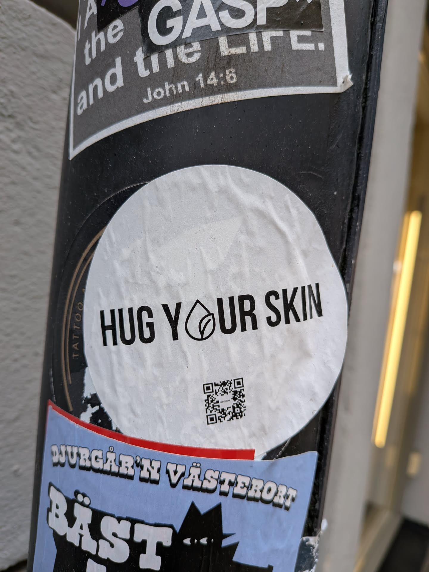 A circular white sticker with bold black text that reads 'HUG YOUR SKIN'. The 'O' in 'YOUR' is replaced by a stylized black logo depicting a drop shape with a leaf inside. Below the text, there is a black and white QR code. The sticker appears slightly wrinkled, indicating its physical presence on a dark pole.