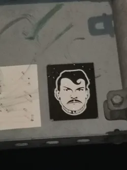 The sticker is rectangular with a black background, featuring a stylized, high-contrast portrait of a man's head and shoulders. The man has a strong jawline, a prominent handlebar mustache, and slicked-back hair with a distinctive curl or pompadour on the forehead. His eyes are narrow and intense, conveying a determined or stern expression. The entire figure is rendered in white outlines and fills against the black background. Around the base of his neck, forming a collar-like shape, is the text "JOE LOUIS" in white capital letters. Small white dots are scattered in the black background, resembling stars or a textured effect.