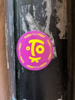 Street sticker A round sticker with the text "Tokyo Film Fest" repeated around the perimeter and a stylized logo in the center. The sticker is purple with yellow text and logo. It's affixed to a dark-colored surface.