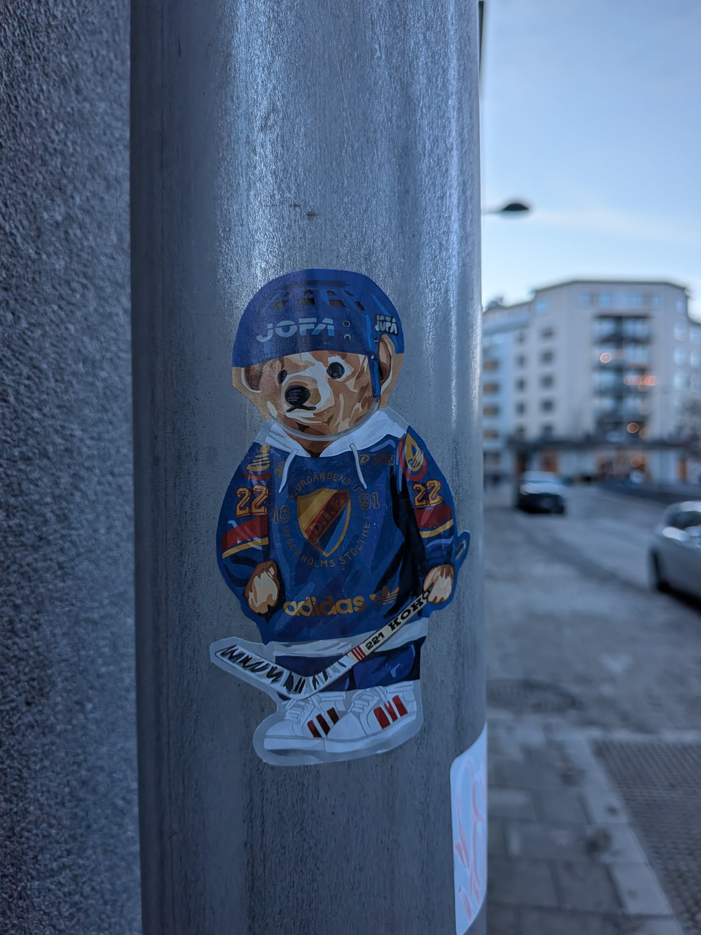A die-cut sticker depicting a brown bear character dressed in full ice hockey gear. The bear wears a blue hockey helmet with 'JOFA' in white text, and a blue hockey jersey with white sleeves and red/yellow accents. The jersey features the Djurgårdens IF crest on the chest, which is a shield design in yellow, red, and blue, surrounded by text 'DJURGÅRDENS IF' and 'STOCKHOLMS STOLTHET'. The number '22' is visible on the right sleeve and '91' on the left. Below the crest, 'adidas' is written in gold. The bear holds a white hockey stick with red and black details, featuring 'KOHO' and '221' text. It wears white sneakers with three red stripes, reminiscent of Adidas Superstars. The sticker has a clean white outline.