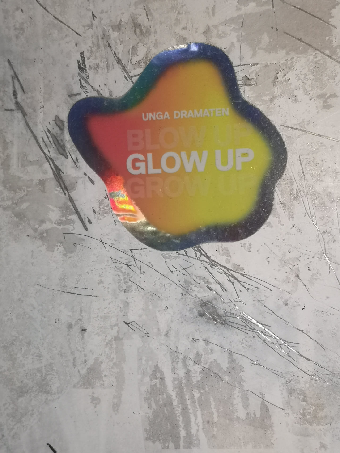 The sticker is an irregularly shaped, organic blob with wavy edges, featuring a striking iridescent or holographic finish. Its color palette transitions smoothly in a gradient from dark blue/purple on the top-left, through green, red, and orange, to bright yellow on the bottom-right. The surface reflects light, creating a rainbow-like sheen in certain areas. Text on the sticker includes 'UNGA DRAMATEN' in a smaller, sans-serif font at the top. Below this, the phrases 'BLOW UP', 'GLOW UP', and 'GROW UP' are stacked vertically in a larger, bold, sans-serif typeface. 'BLOW UP' appears in a reddish-orange, 'GLOW UP' in white, and 'GROW UP' in yellow, blending with the background gradient. The sticker is affixed to a heavily scratched and worn light grey surface.