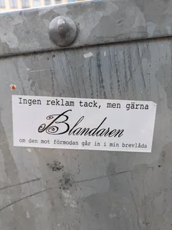 Street sticker A white rectangular sticker is affixed to a gray metal surface. The sticker has black text in a serif font. The text reads "Ingen reklam tack, men gärna Blandaren om den mot förmodan går in i min brevlåda".