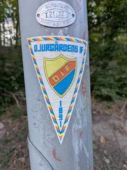 Street sticker The sticker is triangular in shape with a white background. It features a crest with yellow and blue sections divided by a red band. Within the crest are the letters 'D.I.F.' in red. The outer edge of the sticker has a border with a repeating pattern of red, yellow, and blue diagonal lines, enclosed by a thin black outline. At the bottom of the sticker, oriented vertically, are the numbers '1 8 9 1' in blue.