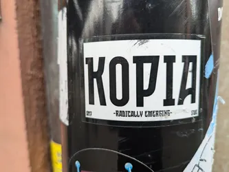 Street sticker A black and white sticker with the word "KOPIA" in bold, uppercase letters. Below the word, it says "-Radically Emerging-". The sticker is attached to a black cylindrical object, possibly a pole or a pipe. 