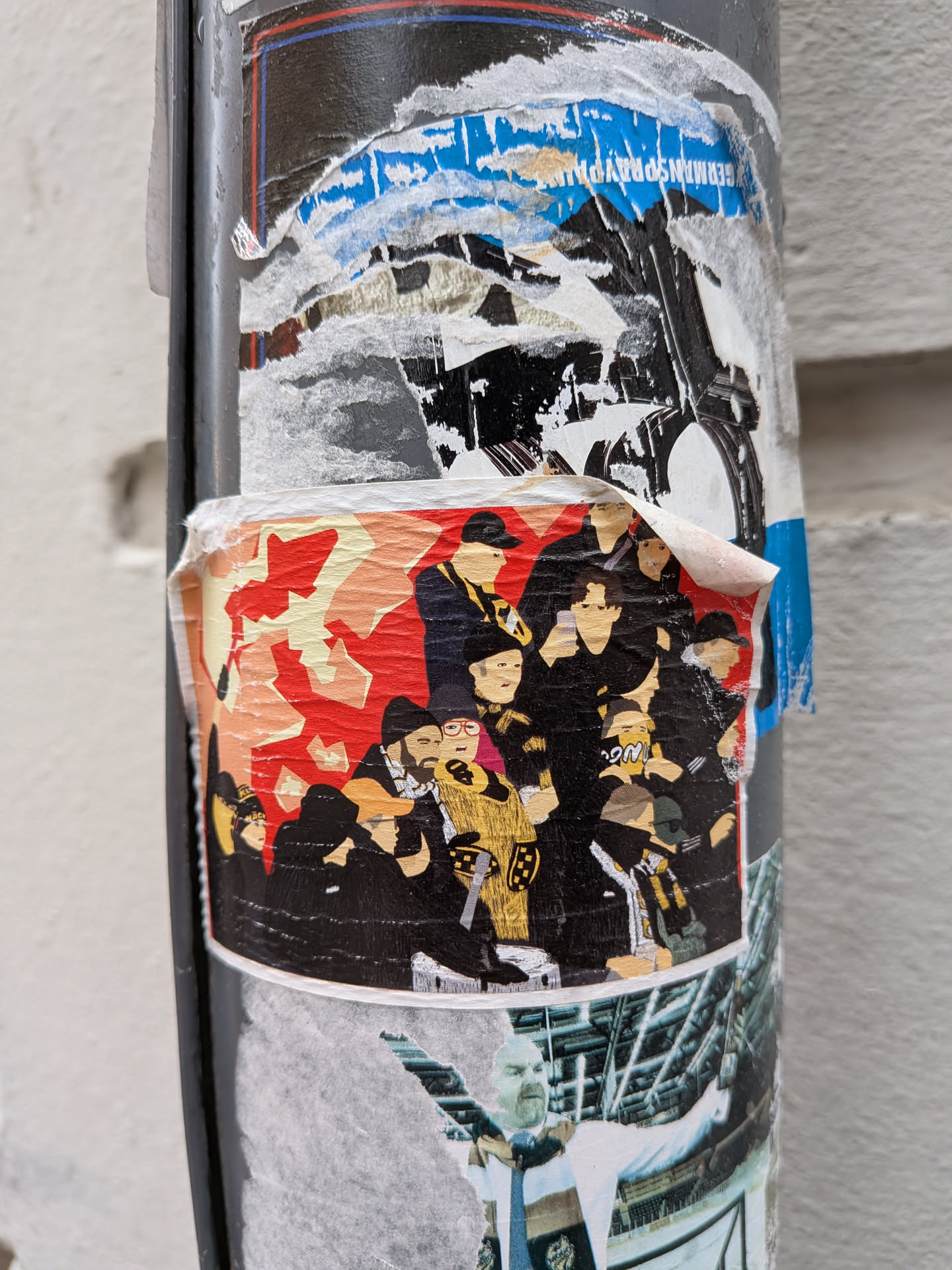 The central sticker features a dense group of stylized human figures, rendered primarily in black, against a dynamic, abstract background composed of red and yellow/beige shapes. Many of the figures wear hats or caps, and some are adorned with yellow or gold-colored scarves or clothing details, suggesting a unified group or fan base. One figure is distinctly wearing glasses. The art style is graphic and somewhat minimalist, focusing on silhouettes and block colors. The sticker itself shows signs of wear, with slightly peeling edges, indicating its outdoor placement on a pole.