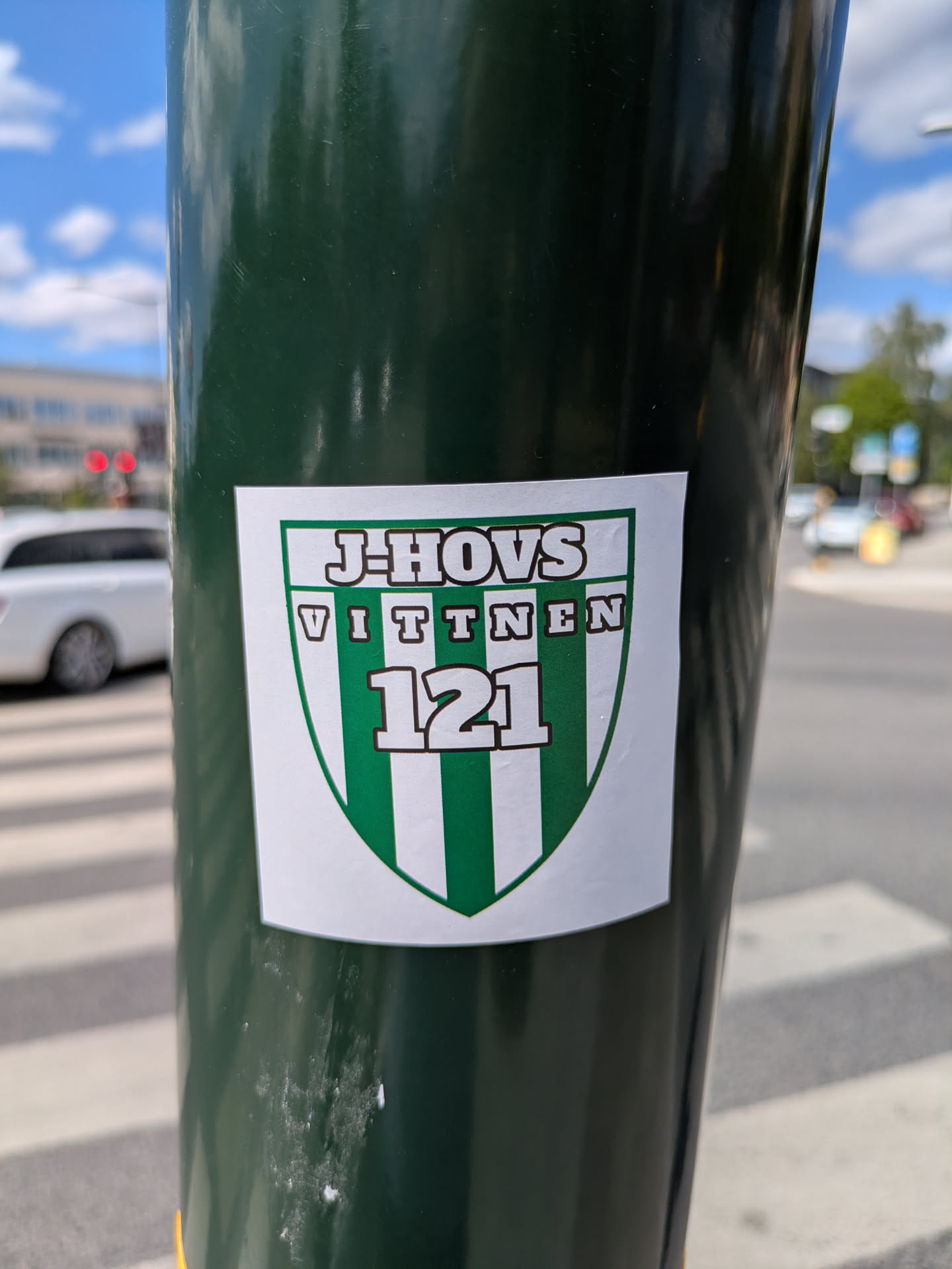 A shield-shaped sticker with a green and white striped design is affixed to a green pole. The sticker reads J-HOVS VITTNEN 121 in bold, capitalized lettering.