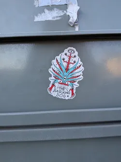 The sticker features a stylized, somewhat monstrous or mythical creature with a predominantly red and light blue color scheme, outlined in black. It has an irregular, die-cut shape with a white border. The central part of the creature's 'face' has a large, prominent eye in the middle, surrounded by several smaller red eyes. Spiky, leaf-like elements in light blue and red radiate outwards from the central mass. Below the main body, two red, claw-like appendages extend downwards. Above the central eye, a tree-like structure with a single large eye at its top emerges, featuring small black dots and spiky elements. The overall style is illustrative and whimsical.