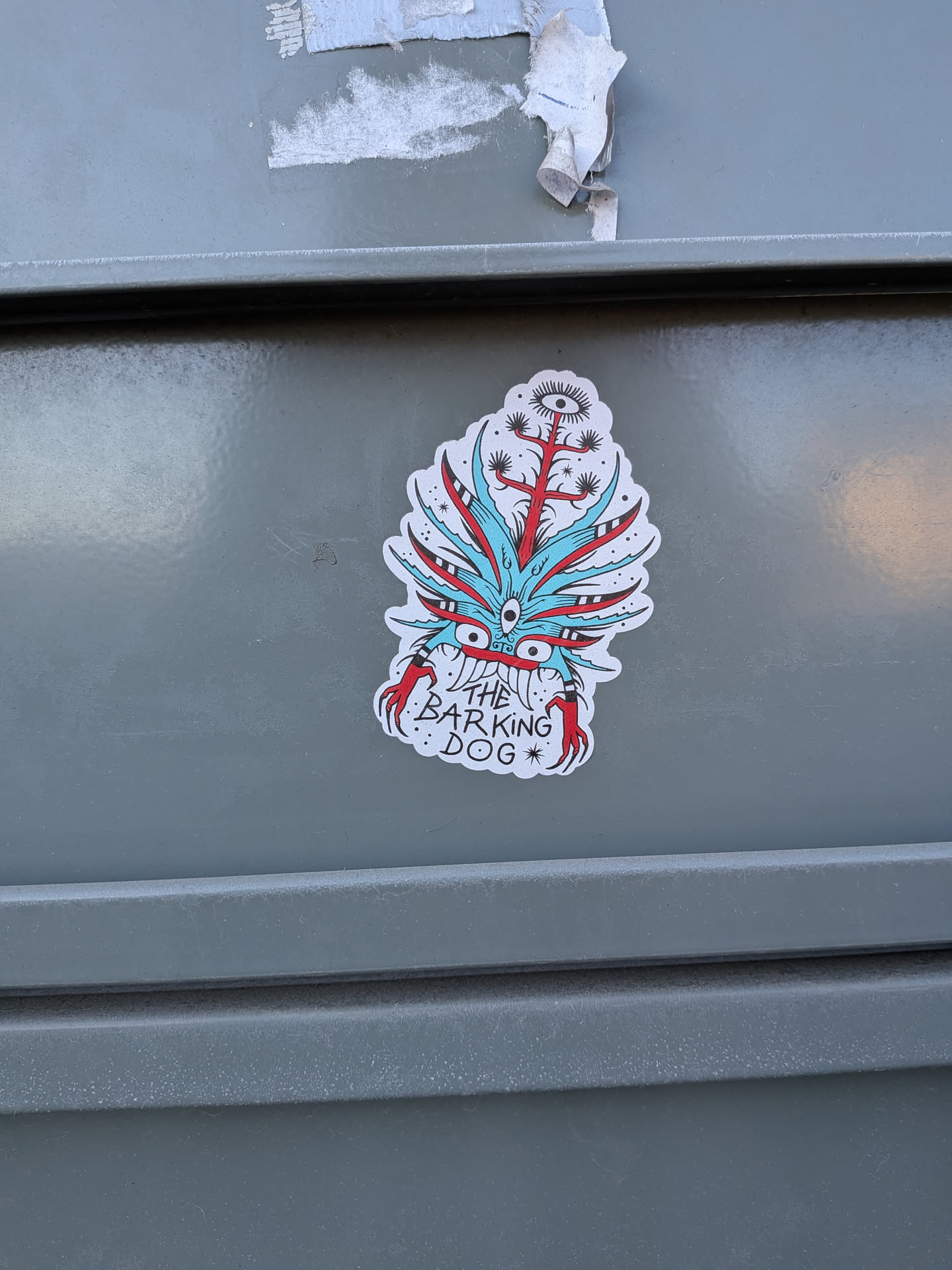 Street sticker The sticker features a stylized, somewhat monstrous or mythical creature with a predominantly red and light blue color scheme, outlined in black. It has an irregular, die-cut shape with a white border. The central part of the creature's 'face' has a large, prominent eye in the middle, surrounded by several smaller red eyes. Spiky, leaf-like elements in light blue and red radiate outwards from the central mass. Below the main body, two red, claw-like appendages extend downwards. Above the central eye, a tree-like structure with a single large eye at its top emerges, featuring small black dots and spiky elements. The overall style is illustrative and whimsical.