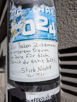 The central sticker is a white rectangular piece of paper with handwritten text in black ink. It is slightly wrinkled and appears to be adhered to a metal pole. Below the main text, a faint 'MONTANA CANS' logo is visible in gray. This white sticker partially covers an older, blue sticker with white text that includes 'MARE!' and '24', which is torn and worn, revealing some yellow patterns underneath. The overall appearance suggests a raw, personal message placed in an urban environment.