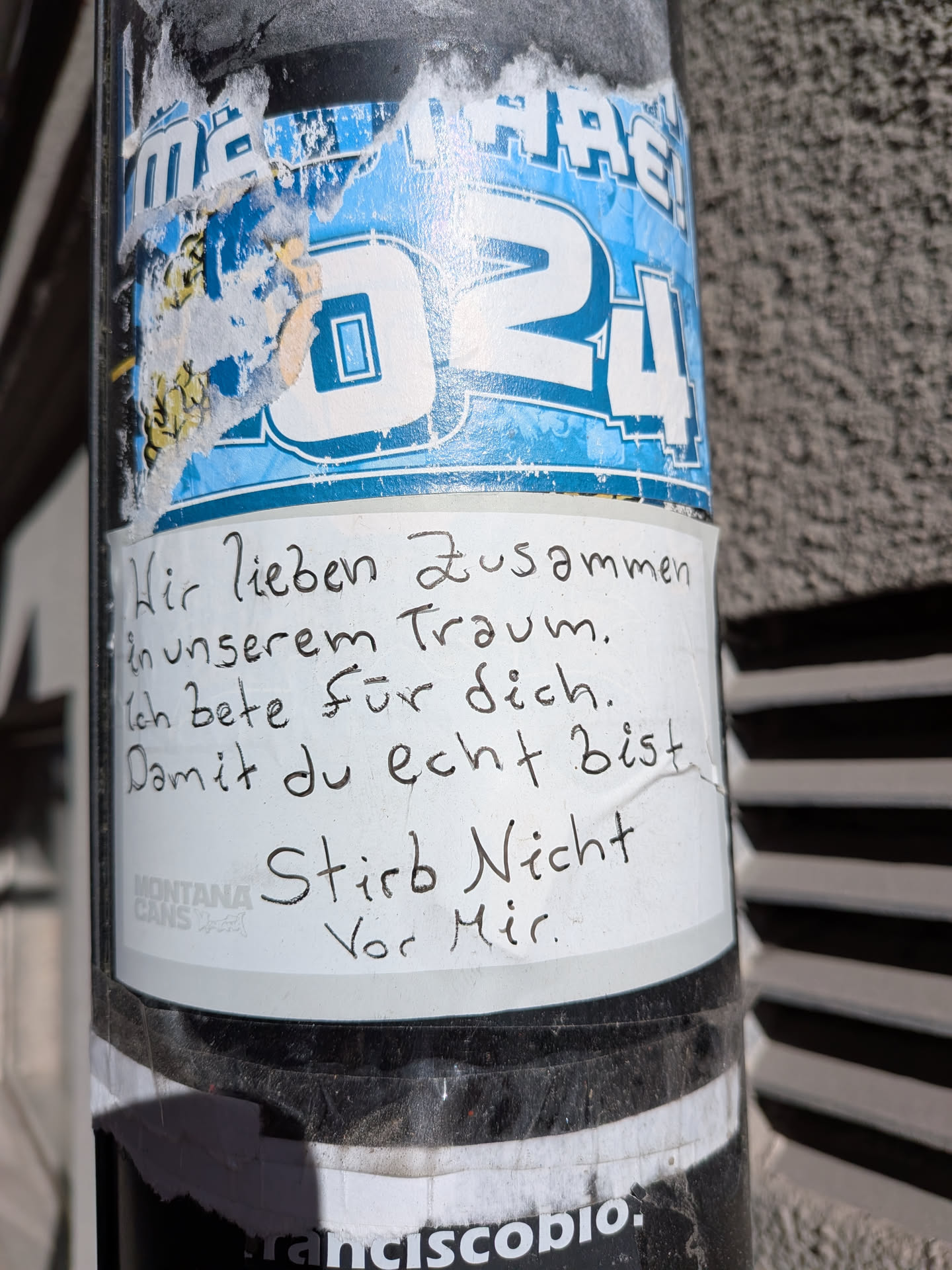 The central sticker is a white rectangular piece of paper with handwritten text in black ink. It is slightly wrinkled and appears to be adhered to a metal pole. Below the main text, a faint 'MONTANA CANS' logo is visible in gray. This white sticker partially covers an older, blue sticker with white text that includes 'MARE!' and '24', which is torn and worn, revealing some yellow patterns underneath. The overall appearance suggests a raw, personal message placed in an urban environment.