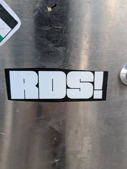 Street sticker A rectangular sticker with a solid black background and slightly rounded corners. The sticker features the text "RDS!" in large, bold, white, sans-serif block letters. The contrast between the white text and the black background is stark, making the text highly prominent. The sticker is applied to a brushed metal surface, showing some wear and tear around its edges.